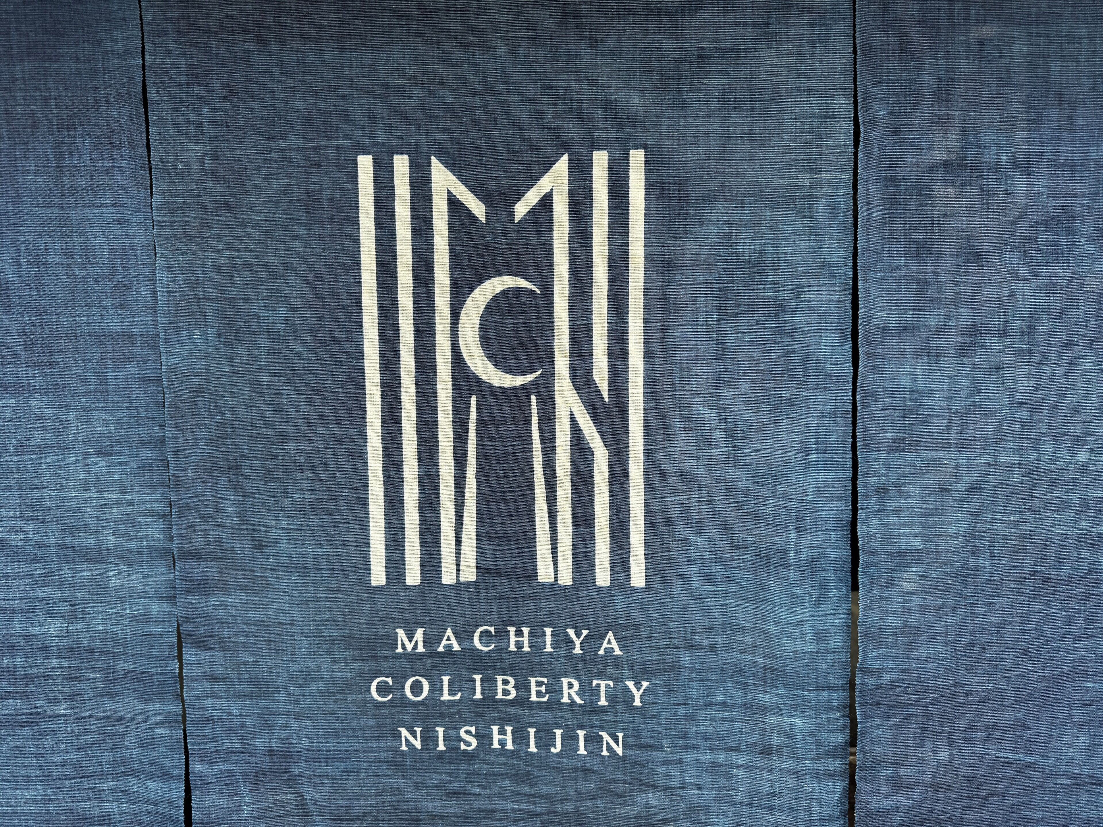 Un logo avec le texte « MACHIYA COLIBERTY NISHIIN » sur une surface texturée.