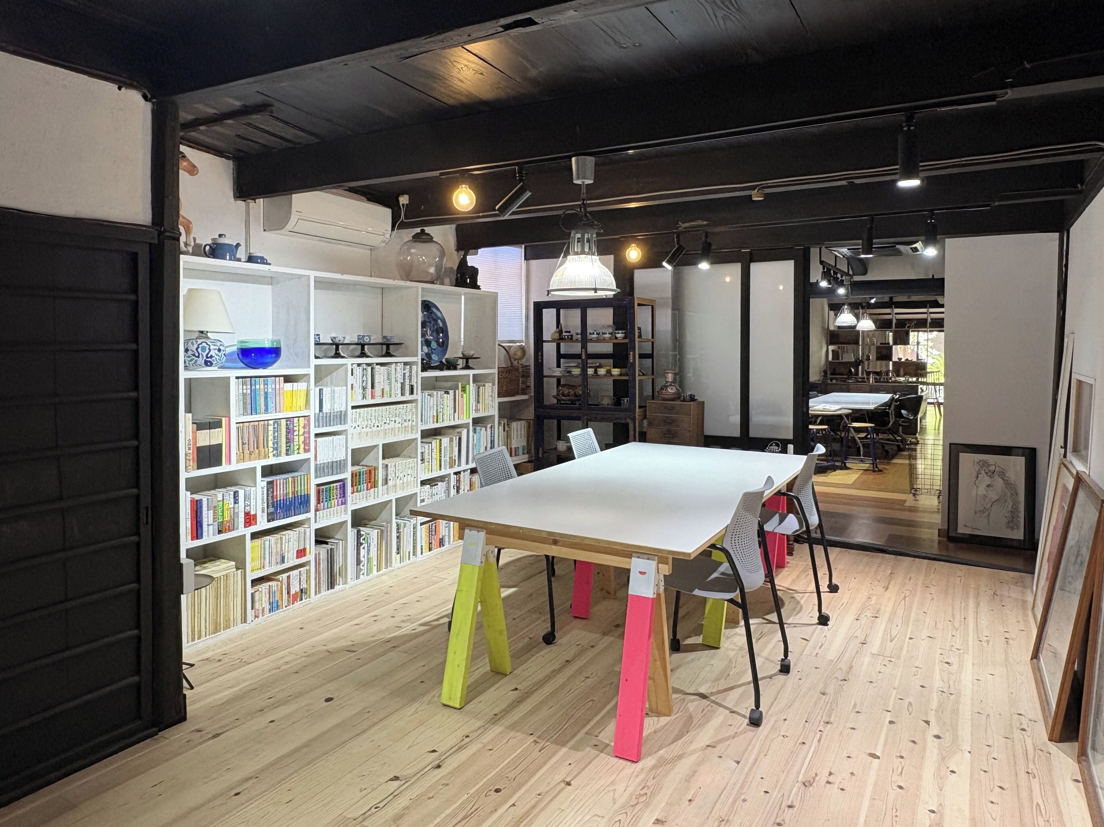 Ein Zimmer mit Holzboden, einem weißen Tisch mit bunten Stühlen, einem Bücherregal voller Bücher und einem gerahmten Bild an der Wand.