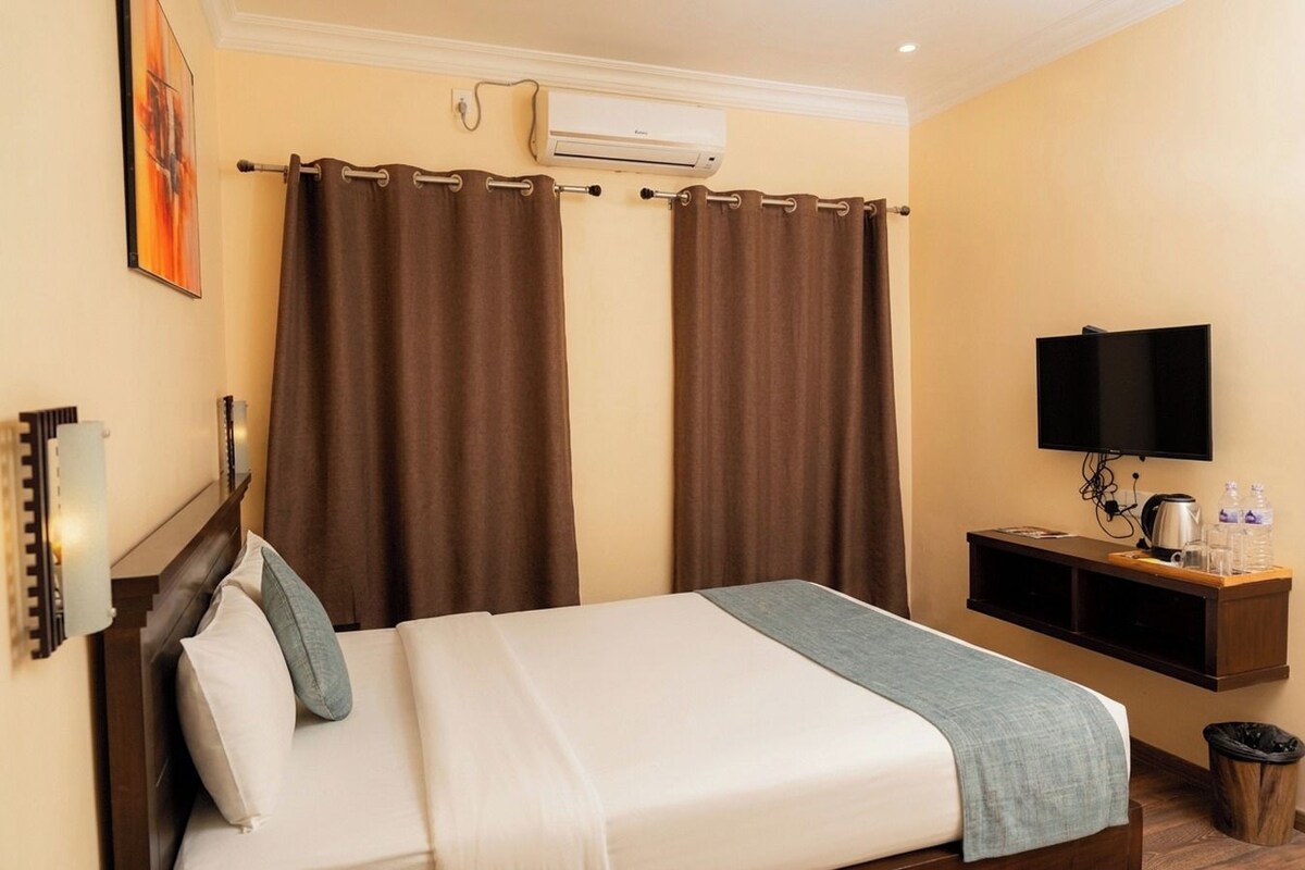 Habitación de hotel con cama, cortinas, televisión, una mesita con una tetera y un cubo de basura.