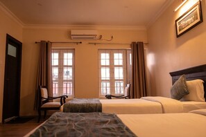 A hotel room with two beds, a chair, a window with curtains, an air conditioning unit, and a painting on the wall.
