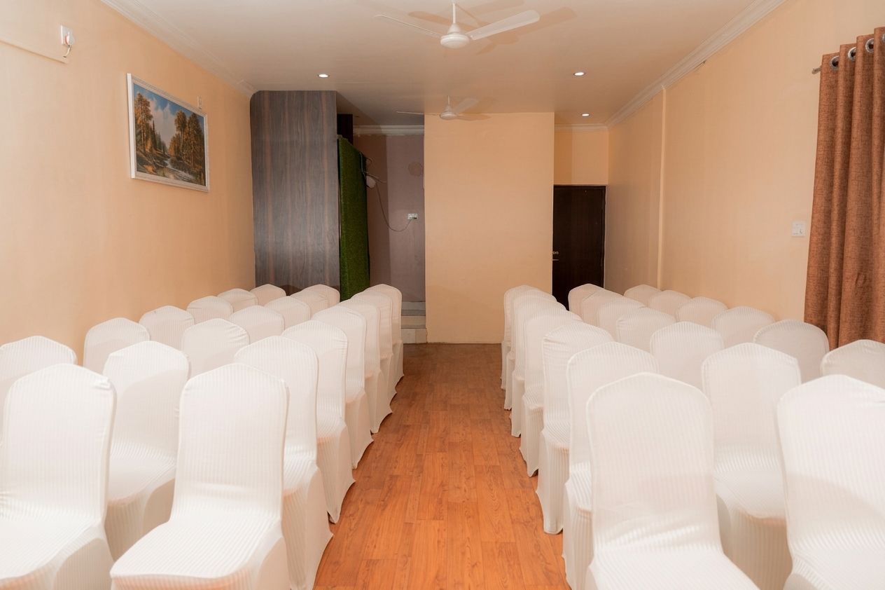Une salle de conférence avec des rangées de chaises blanches, un plancher en bois, un ventilateur au plafond et un tableau accroché au mur.
