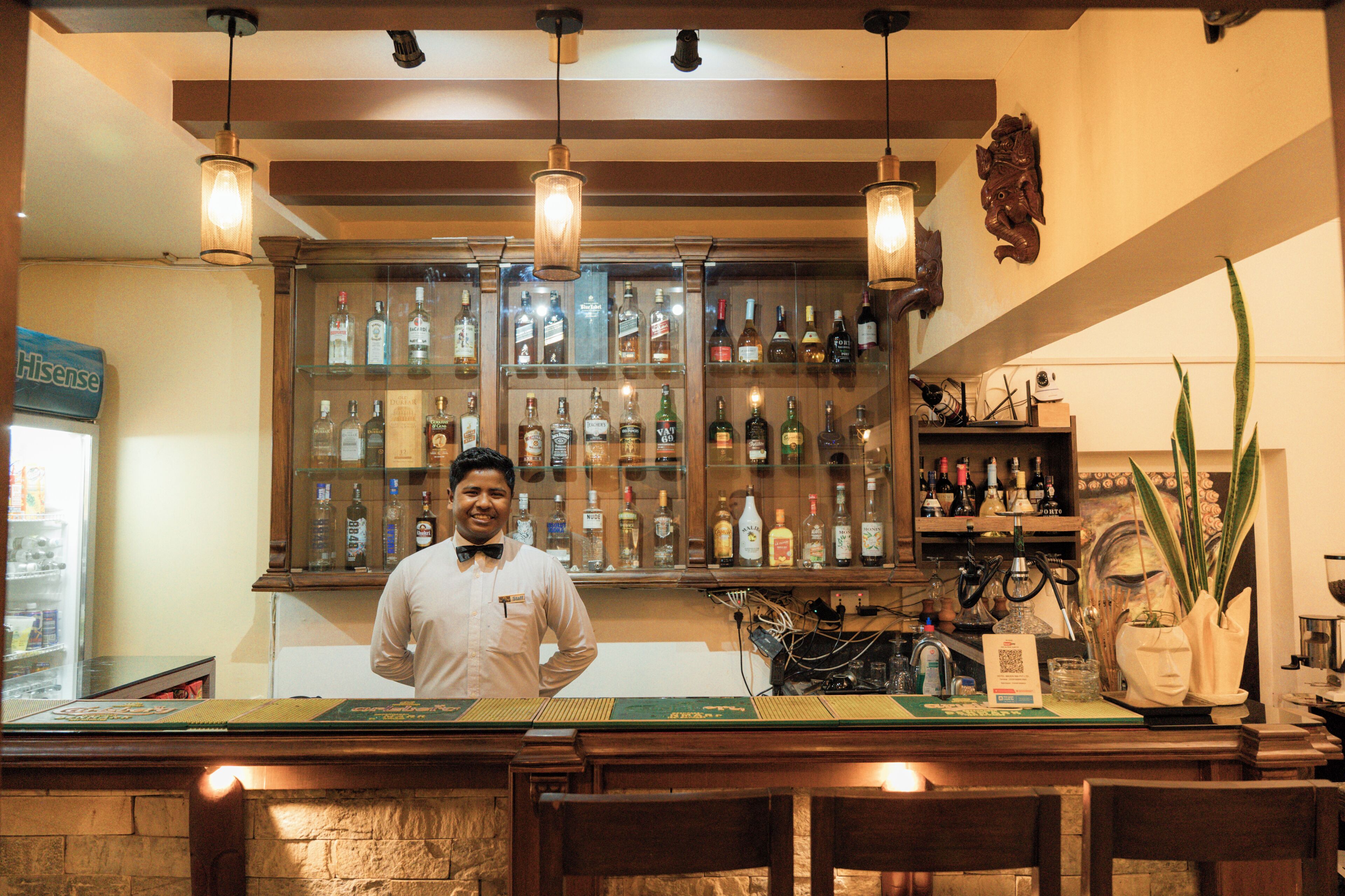 Zona de bar con un cantinero, estantes llenos de botellas de licores, luces colgantes y un elemento decorativo en la pared.