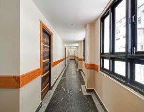 A long, narrow hallway with a wooden door on the left and large windows on the right. - Premium Room Mantto Plaza (Kanpur)