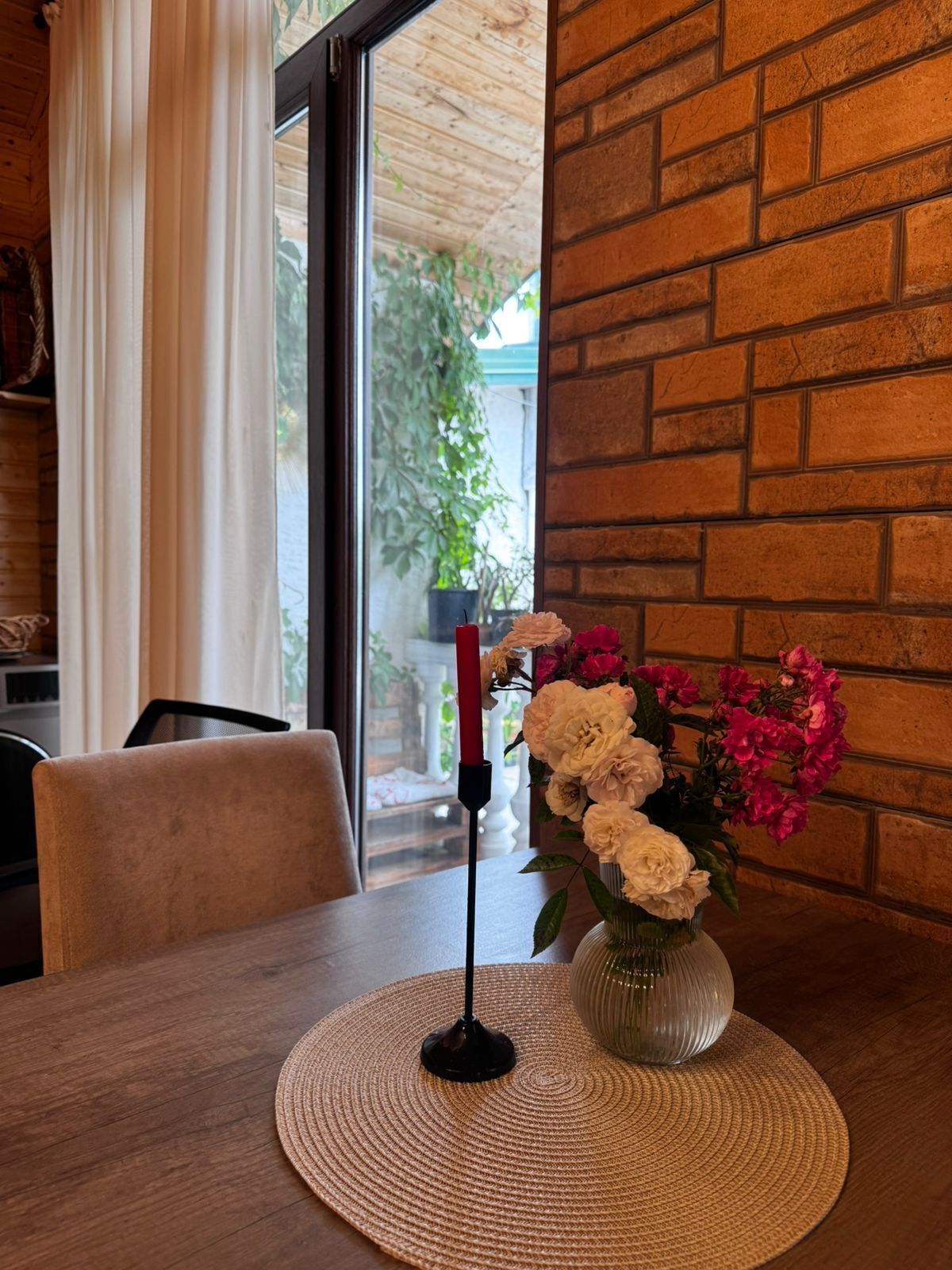 Une table à manger agrémentée d’un vase de fleurs et d’une bougie, placée devant un mur de briques et une fenêtre ornée de rideaux.
