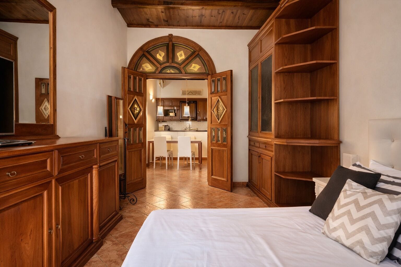Habitación de hotel con cama, armario, escritorio y una cocina visible a través de una puerta abierta.