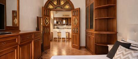 Habitación de hotel con cama, armario, escritorio y una cocina visible a través de una puerta abierta.
