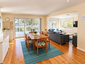 A spacious living area with a dining table, sofa, and a balcony with a view. - Wirani on the Water (Sanctuary Point)
