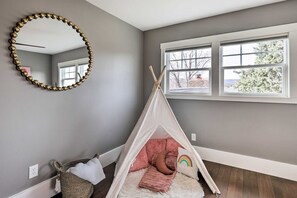 Ein Zimmer mit grauer Wand, weißer Decke, einem großen Spiegel, einem Fenster mit Blick auf Bäume und einem Zelt in Form eines Tipis mit einem Regenbogenkissen.