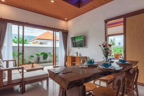 Zona pranzo con un grande tavolo di legno, sedie e vista su una casa attraverso una porta scorrevole in vetro.