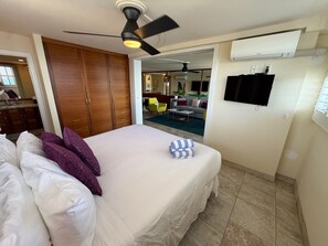 A hotel room with a bed, a ceiling fan, an air conditioning unit, a television, and a view into the adjacent living area. - Oceanview 2BR/2BA condo with pool, AC, balcony, and beach access-Kanai A Nalu 317 (Wailuku)