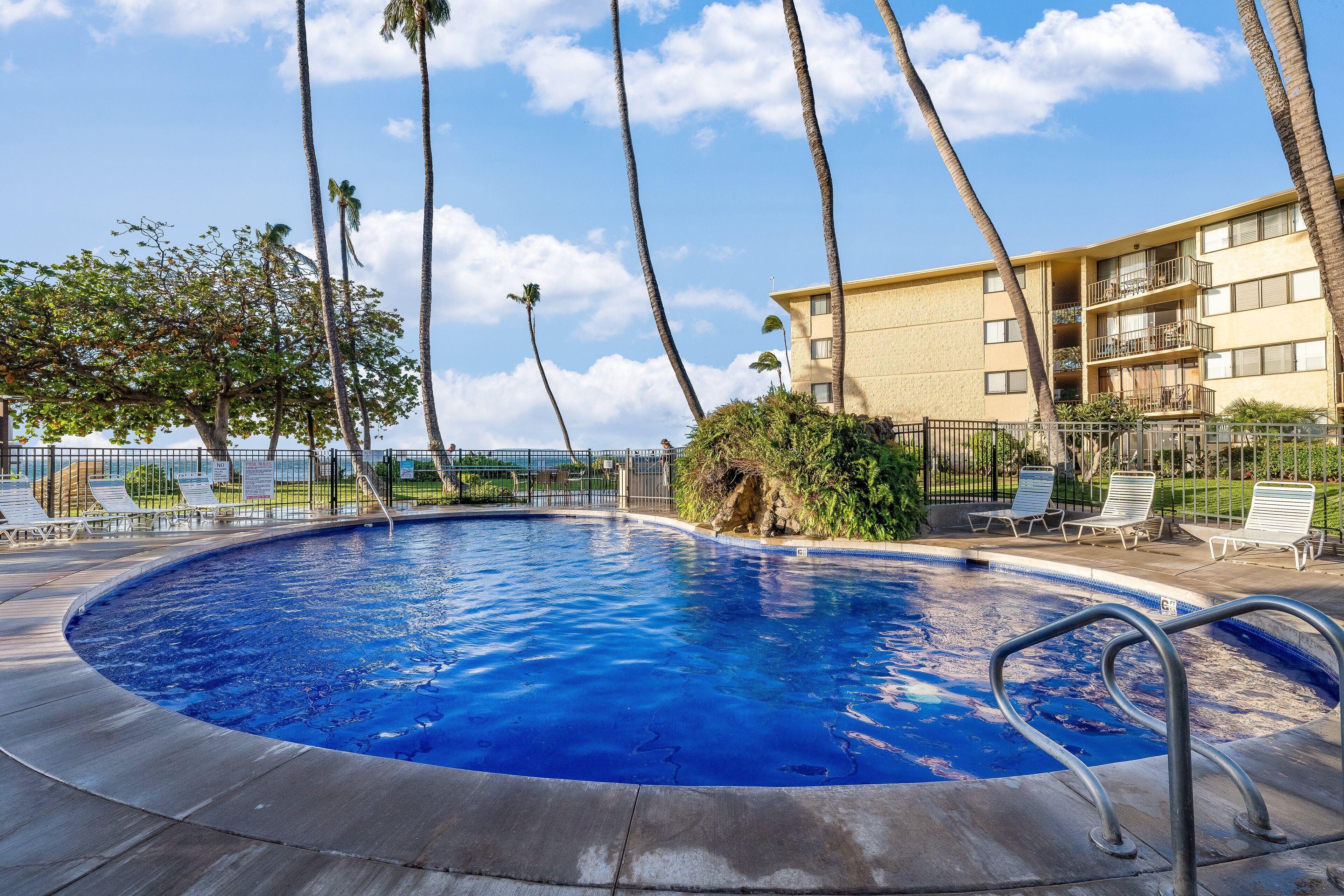 A swimming pool with a view of the ocean, surrounded by palm trees and a building with balconies.