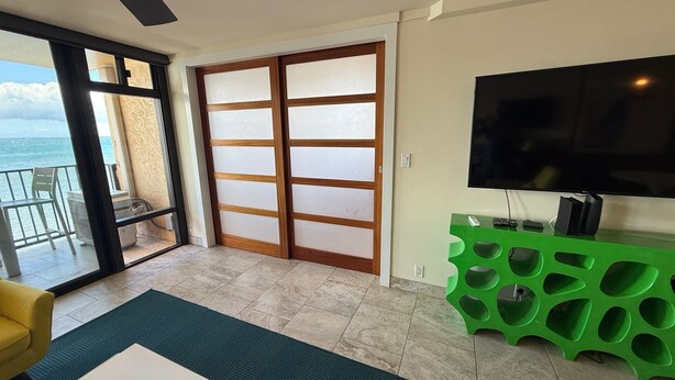 A living room with a sliding glass door leading to a balcony, a flat-screen TV mounted on the wall, and a green entertainment unit. - Oceanview 2BR/2BA condo with pool, AC, balcony, and beach access-Kanai A Nalu 317 (Wailuku)
