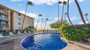 A swimming pool with lounge chairs and palm trees, surrounded by a building with balconies. - Oceanview 2BR/2BA condo with pool, AC, balcony, and beach access-Kanai A Nalu 317 (Wailuku)