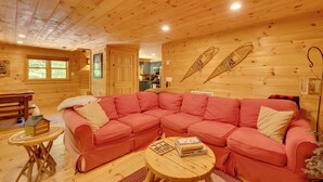 Ein gemütliches Blockhauss-Interieur mit einem großen, pinkfarbenen Ecksofa, Holzdielen und einem durch eine offene Tür erkennbaren Küchenbereich.