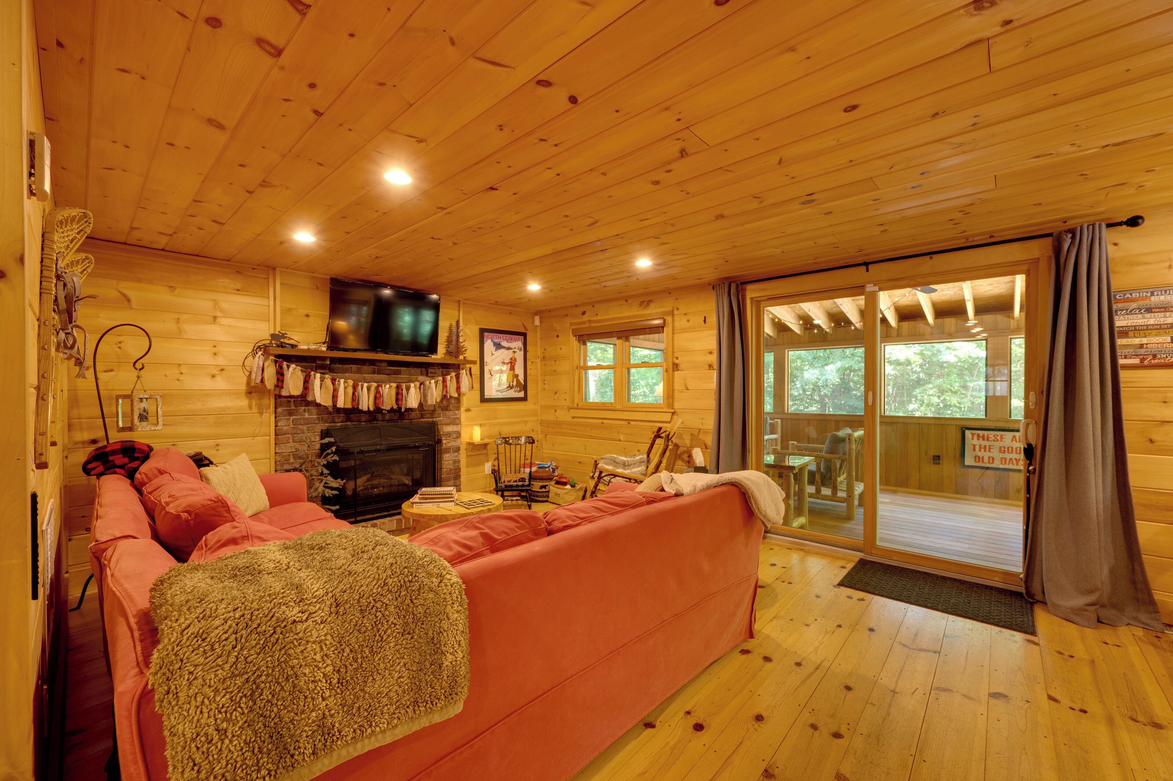 A wooden cabin interior with a fireplace, a television, a sofa, and a sliding glass door leading outside.
