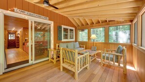 Ein gemütliches Blockhaus-Interieur mit Holzbalkendecke, Deckenventilator, einer Couch, zwei Sesseln, einem Puzzeltisch und einem großen Fenster mit Blick auf den Wald.