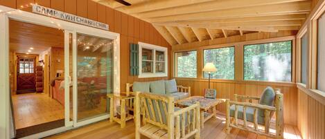 Interior acogedor de una cabaña con techo de madera, ventilador de techo, un sofá, dos sillones, una mesa para rompecabezas y un amplio ventanal con vistas al bosque.