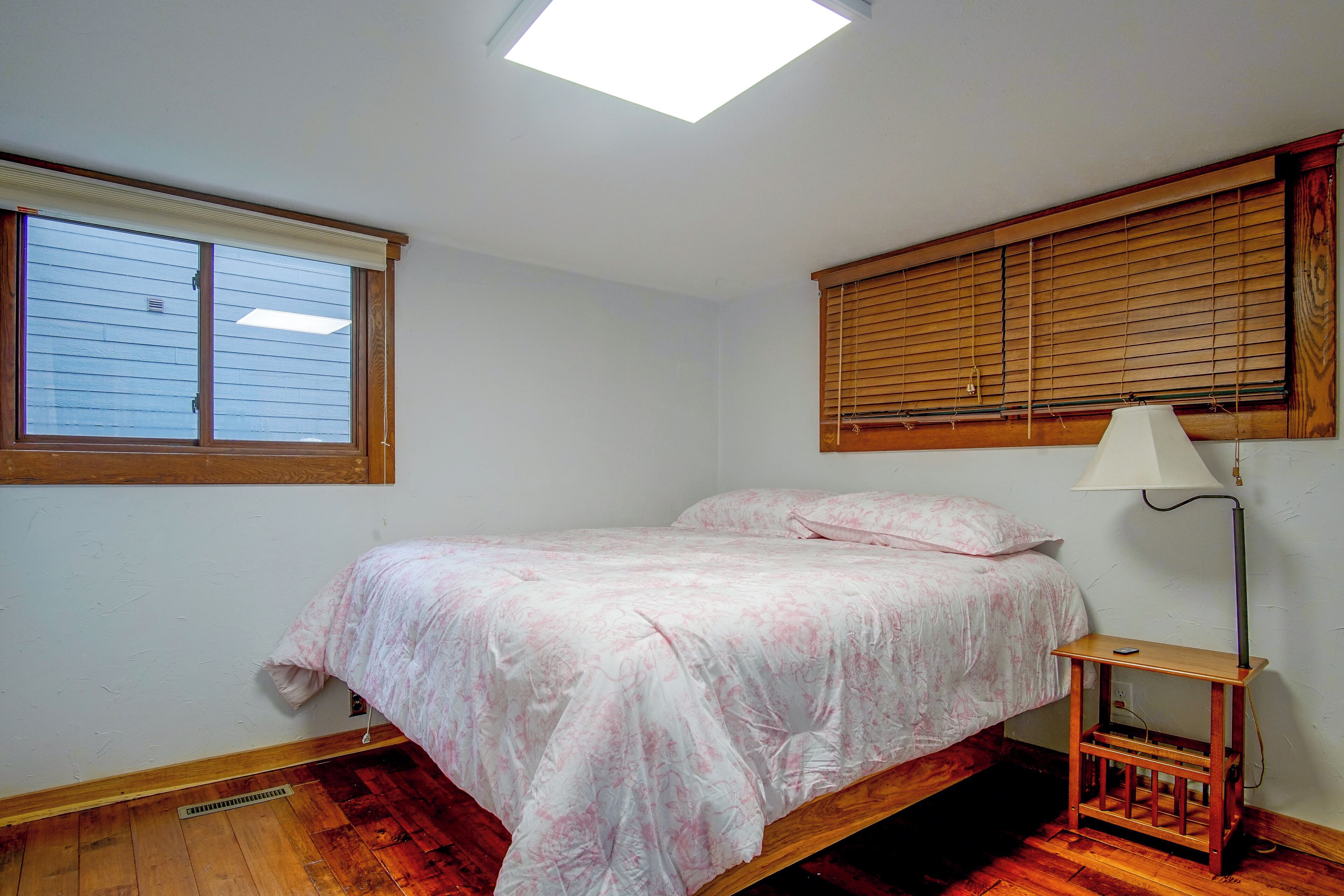 A bedroom with a bed, a nightstand, a lamp, a window with blinds, and a skylight.