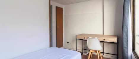 A small, sparsely furnished room with a single bed, a desk, and a chair.