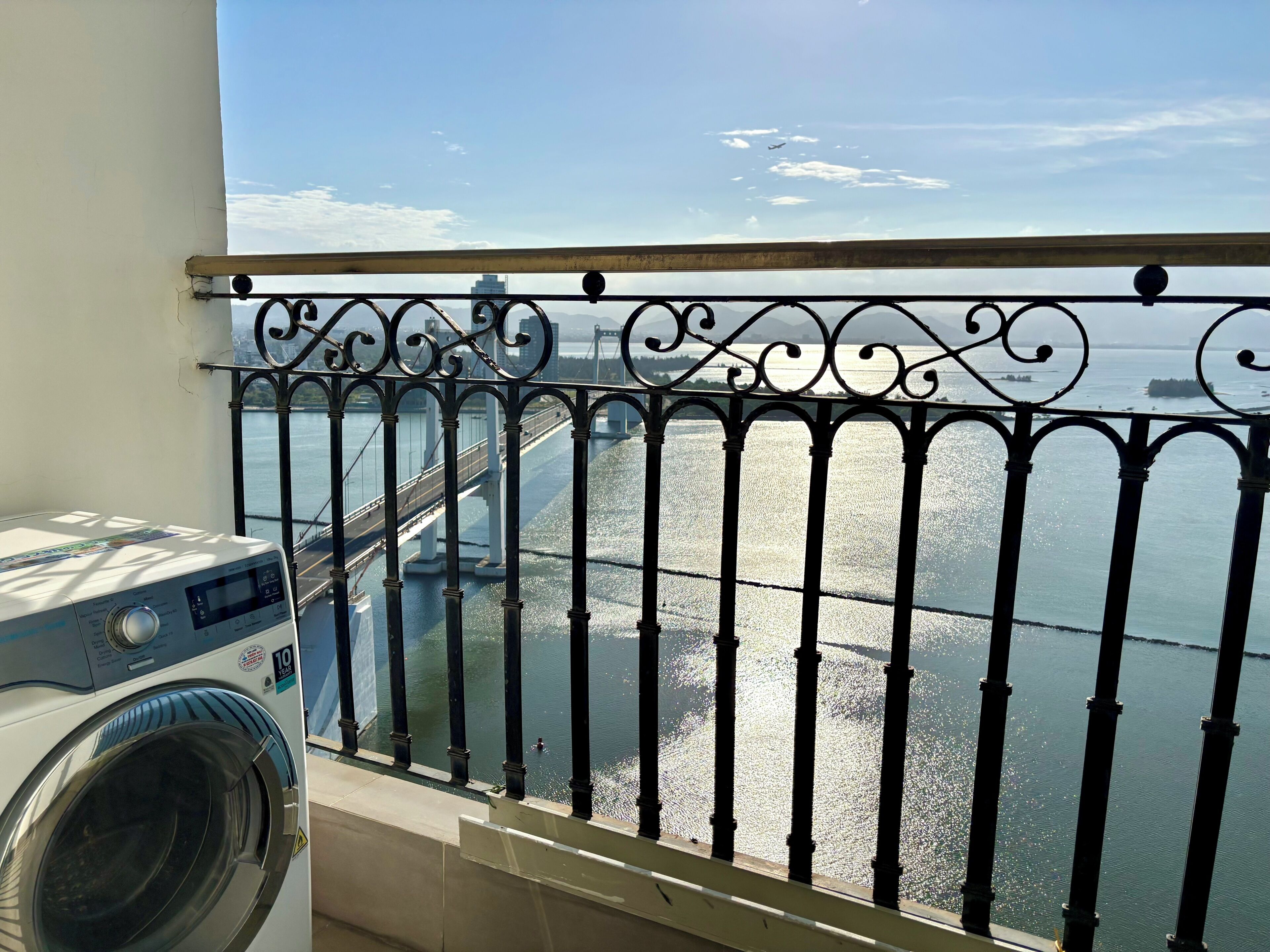 A balcony with a washing machine, a metal railing, and a view of a bridge over water.