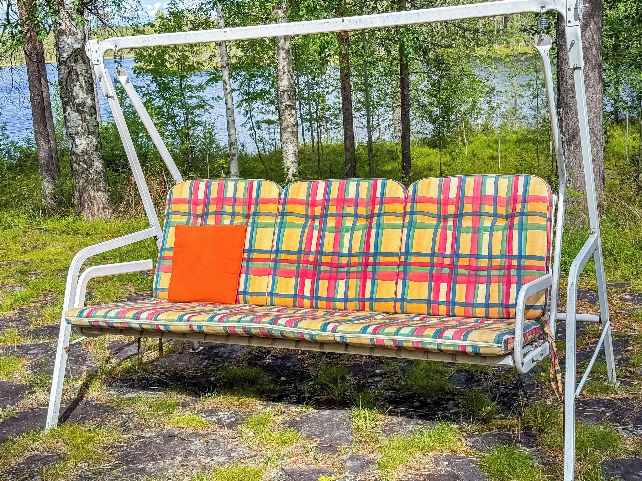 Een metalen schommelzitplaats met een kleurrijke geruit kussen en een oranje kussen, voor een natuurlijke achtergrond met bomen en een waterpartij.