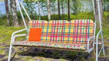 Een metalen schommelzitplaats met een kleurrijke geruit kussen en een oranje kussen, voor een natuurlijke achtergrond met bomen en een waterpartij.