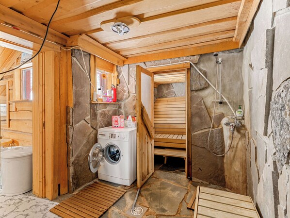 A compact, rustic bathroom with a stone wall, wooden beams, a washing machine, and a sauna.