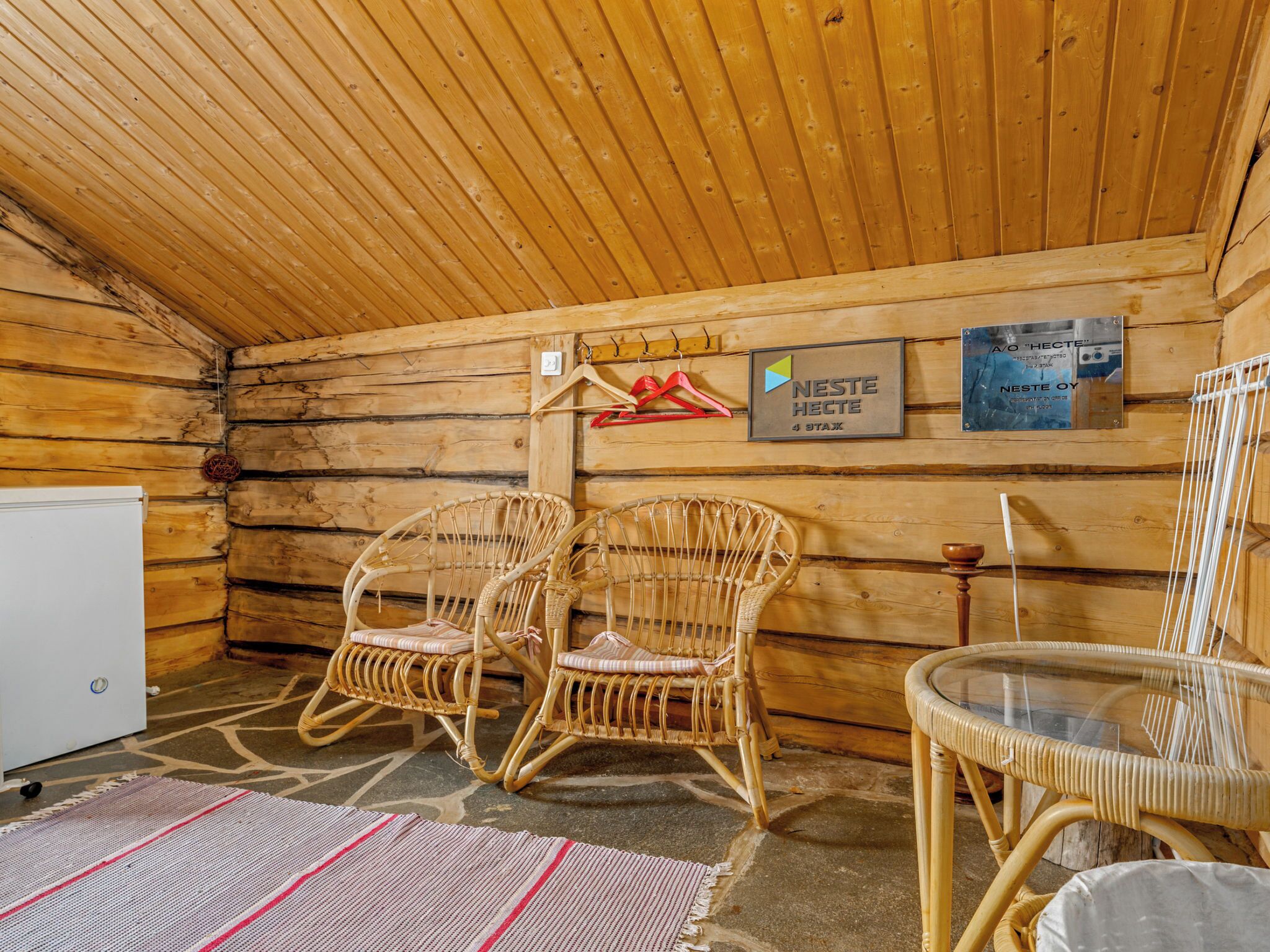 Interior d'una cabana de fusta amb dues cadires de vimbles, una taula de vidre i un frigorífic.