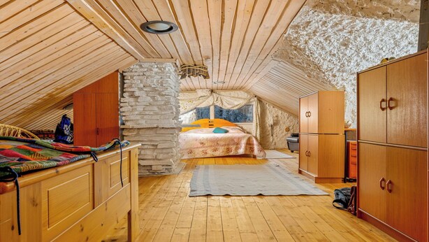 A cozy attic bedroom with a wooden ceiling, a stone wall, a bed with colorful bedding, and wooden furniture.
