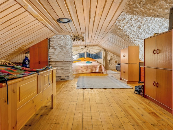 A cozy attic bedroom with a wooden ceiling, a stone wall, a bed with colorful bedding, and wooden furniture.