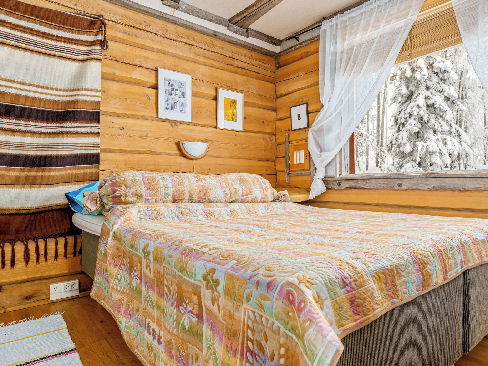 A cozy wooden room with a bed, a window with a view of snow-covered trees, and a wall with framed pictures.