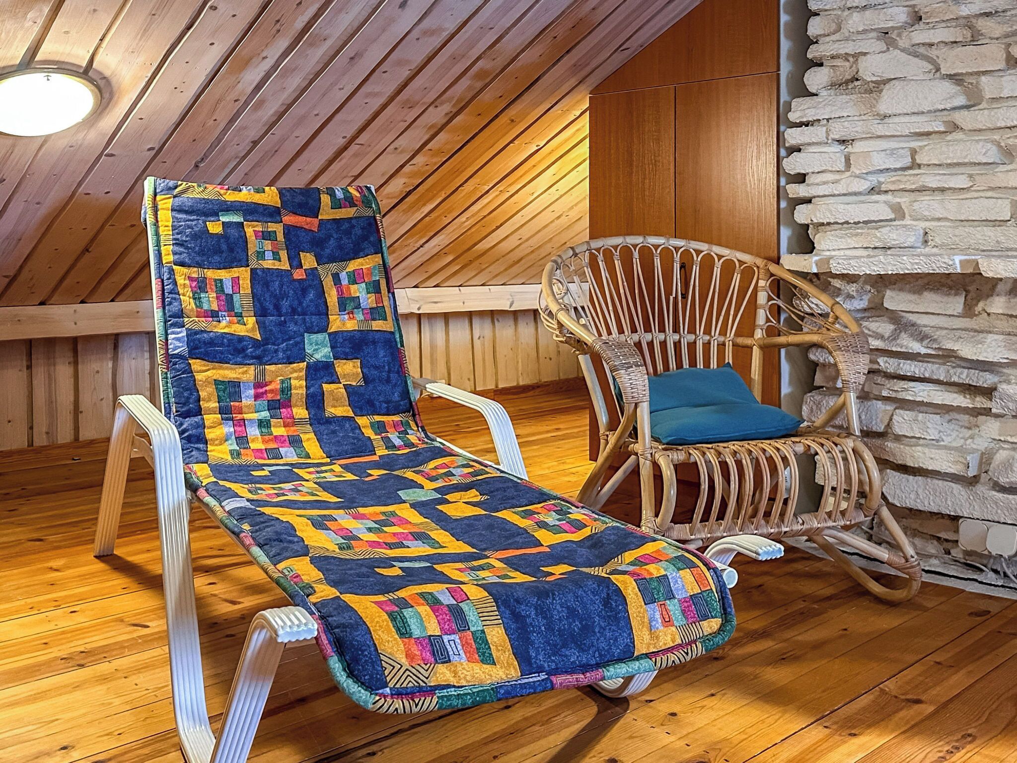 Een houten plafond met dakraam, een houten vloer, een kleurrijke quiltstoel met kussen, en een stenen muur.