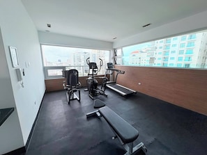 A gym with exercise equipment, a bench, and large windows offering a city view. - Step into elegance and comfort (Santo Domingo)