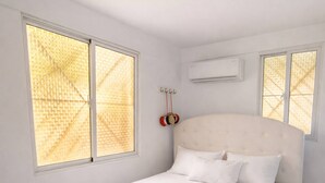 A bedroom with a large bed, two windows with blinds, an air conditioning unit, and a red clock on the wall.