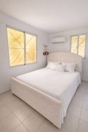 A bedroom with a large bed, two windows with blinds, an air conditioning unit, and a red clock on the wall.