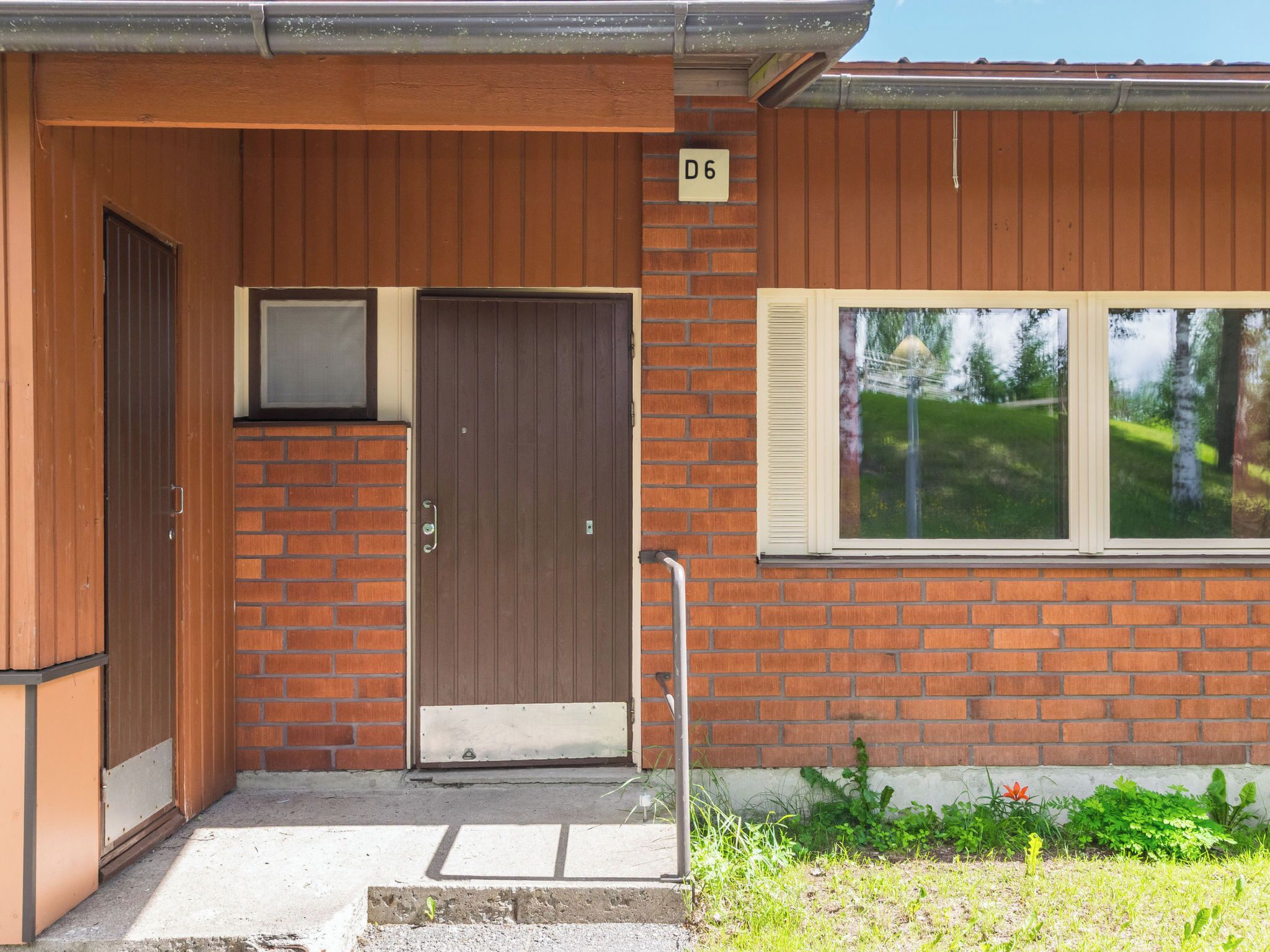 A two-story building with a brown wooden facade, a brown door, and a window with a view of greenery.