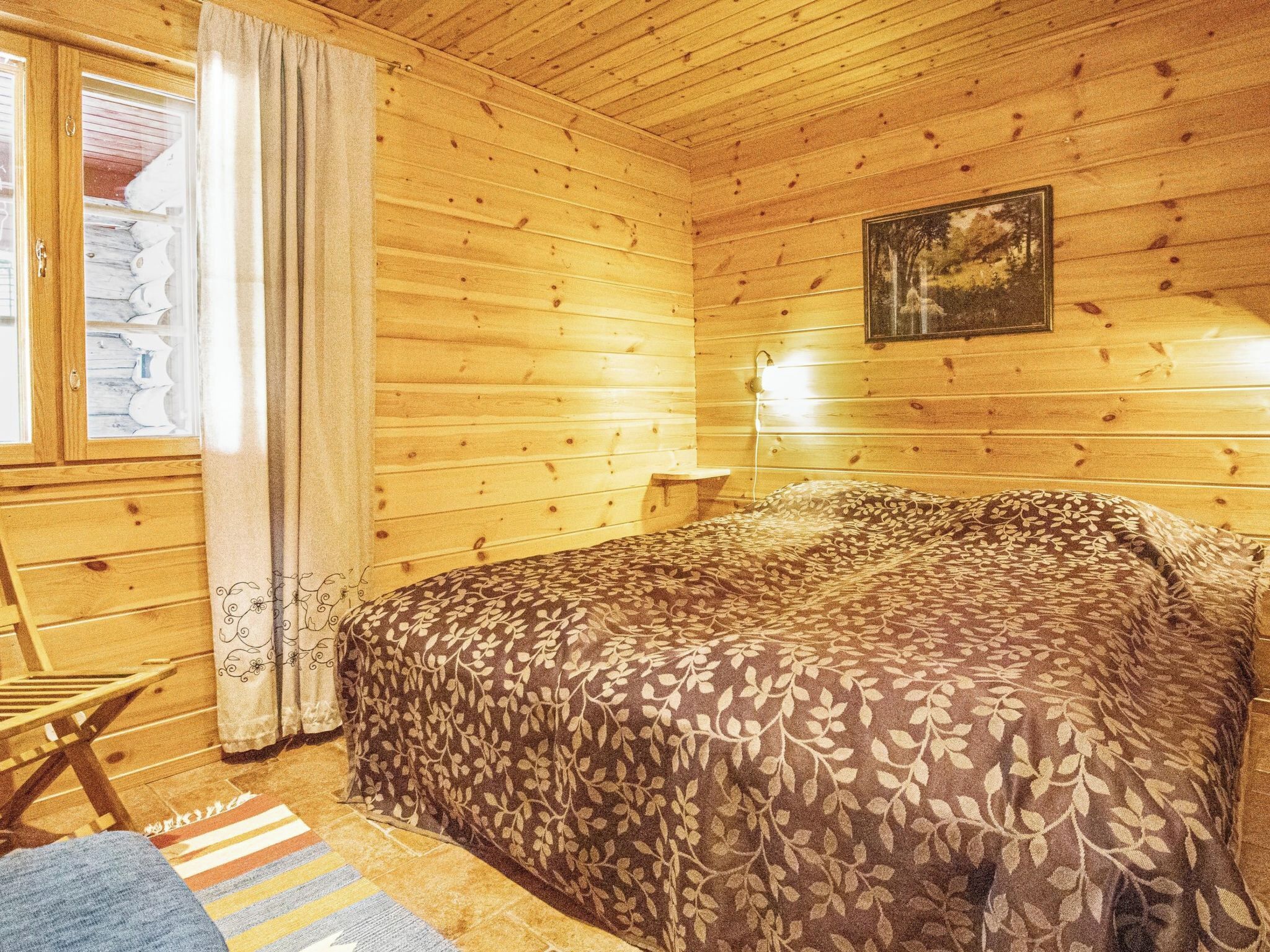 A wooden cabin room with a bed, a small table, and a window with curtains.