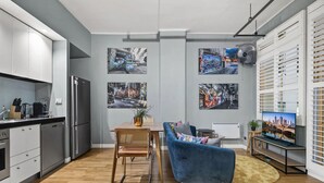 Una cocina moderna con refrigerador empotrado, un área de comedor con sofá azul y una mesa, y un espacio de sala de estar con televisor y obras de arte en las paredes.