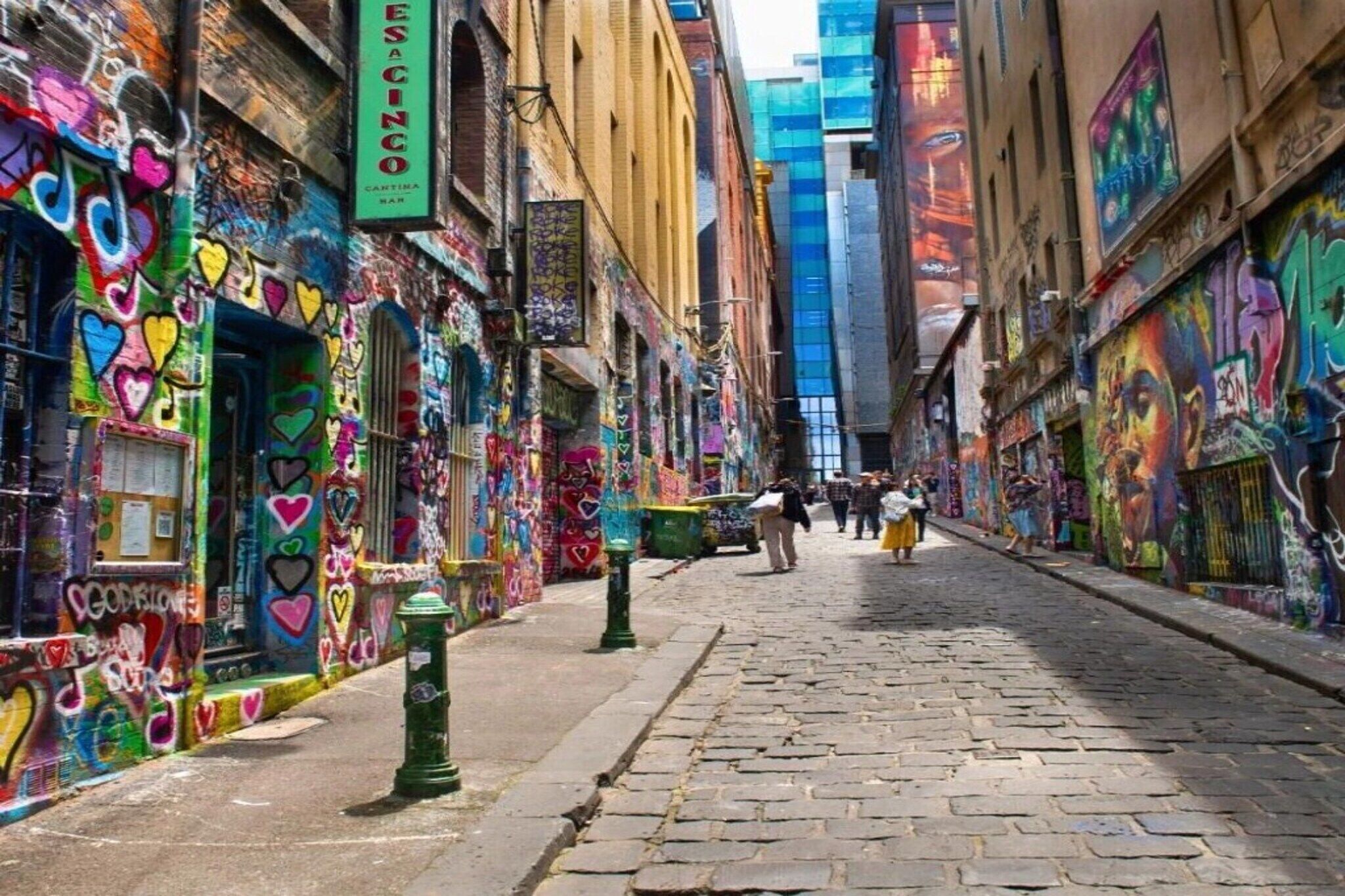 Eine schmale Straße mit an Häusern gesprayten bunten Graffiti, einem sichtbaren Bardekor und einigen Fußgängern.