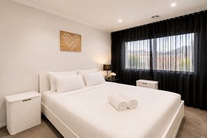 A hotel room with a large bed, a nightstand, a trash bin, a lamp, and a window with curtains. - Spacious 4-Bedroom Family Home in Manor Lakes (Manor Lakes)