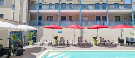 A hotel with a swimming pool, sun loungers, and red umbrellas.