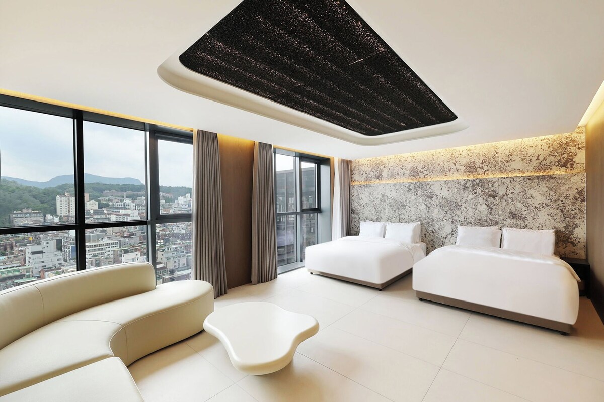 A modern hotel room with a city view, two beds, a sofa, and a unique ceiling light fixture.