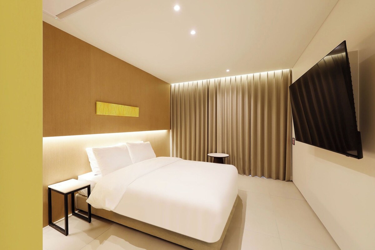 A modern hotel room with a large bed, a bedside table, a small round table, and a flat-screen TV mounted on the wall.
