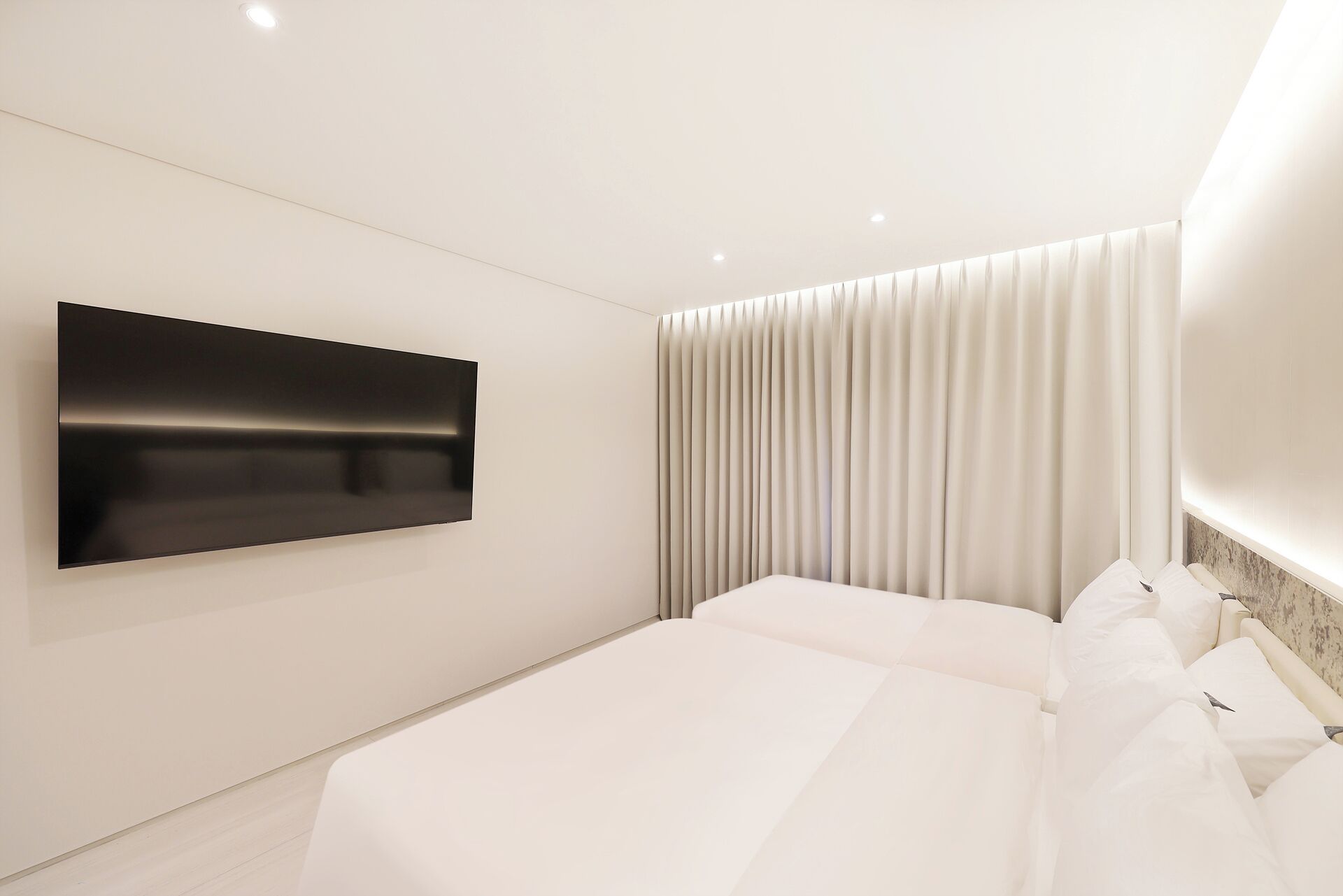 Ein modernes Schlafzimmer mit einem großen Flachbildfernseher an der Wand, einem Bett mit weißer Bettwäsche und einem Vorhang mit vertikalen Falten.