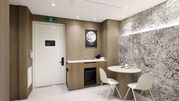 Une chambre d’hôtel moderne avec une porte en bois, une petite kitchenette, une table ronde avec deux chaises et un mur orné d’un motif texturé.