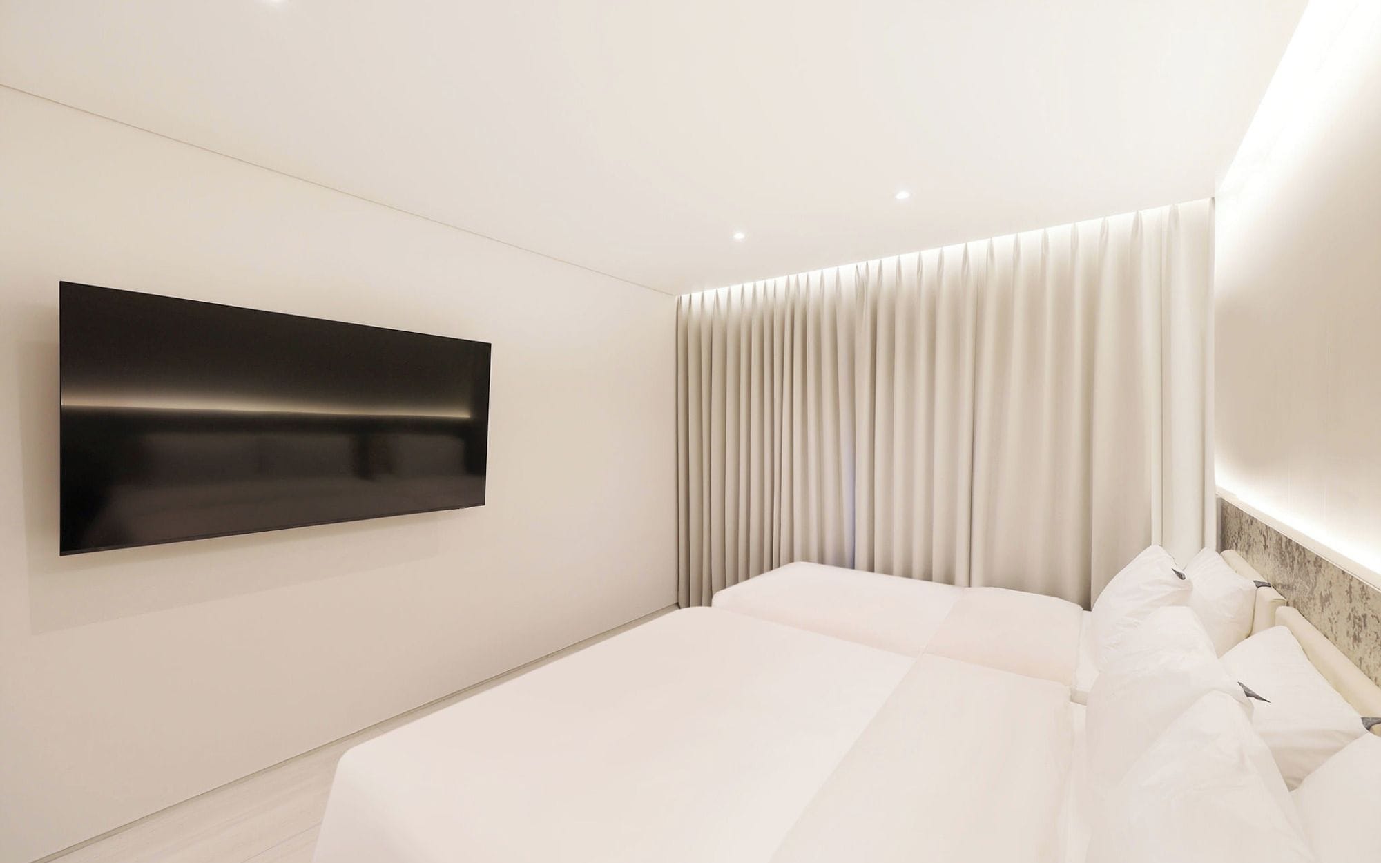 Una habitación de hotel moderna con un televisor de pantalla plana grande montado en la pared, una cama con ropa blanca y un ventanal cubierto por cortinas.