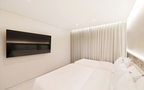 A modern hotel room with a large flat-screen TV mounted on the wall, a bed with white linens, and a curtain-covered window.