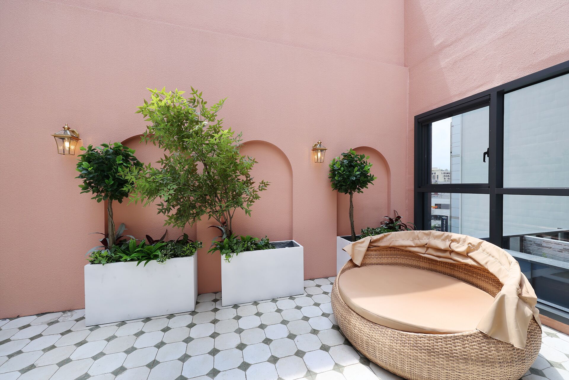 Un balcon doté d’un sol carrelé de carreaux blancs, d’un canapé en osier avec un coussin beige et de plantes en pot disposées contre un mur rose.