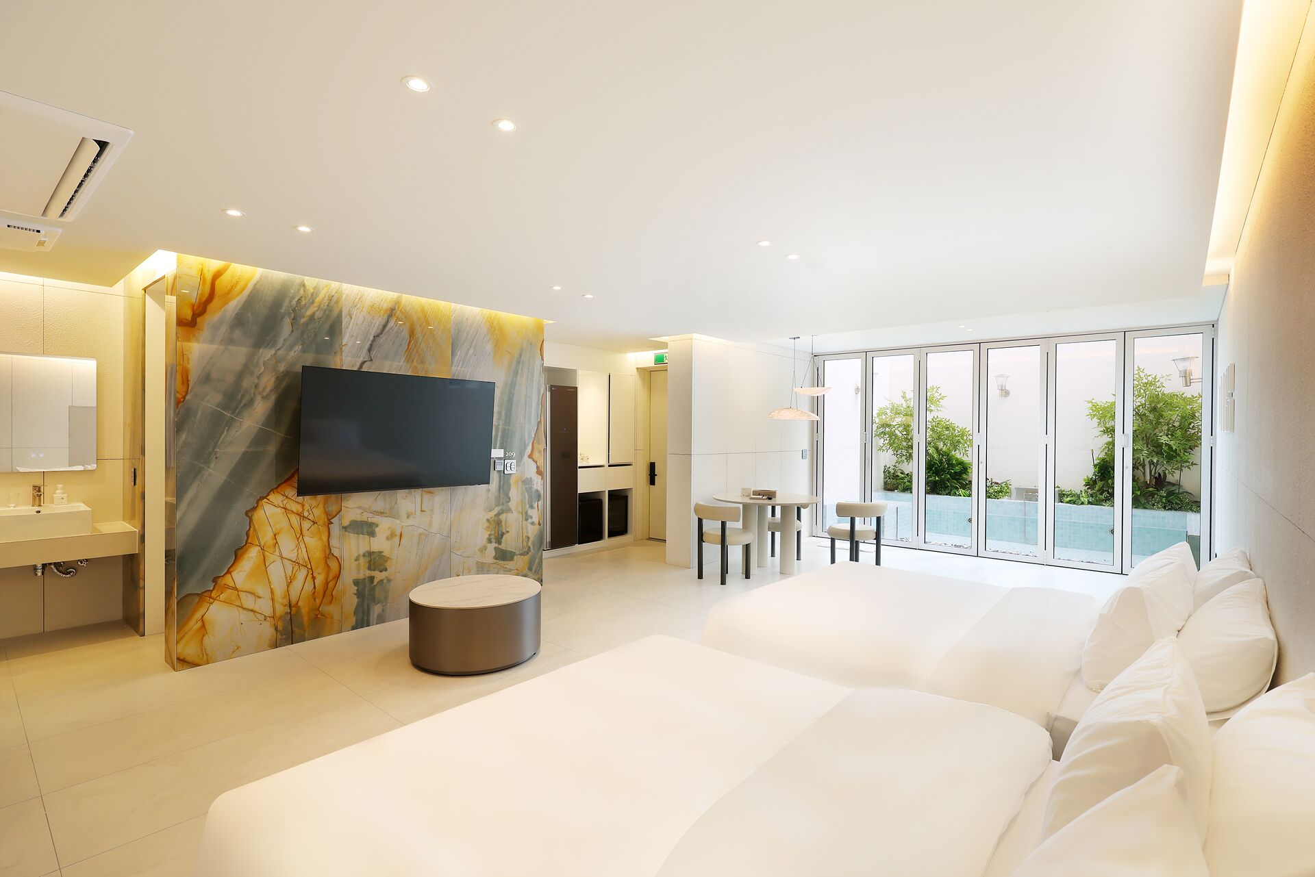 Habitación de hotel moderna con una cama grande, un televisor de pantalla plana montado en una pared de mármol, una mesita redonda con sillas y un balcón con vistas a zonas verdes.