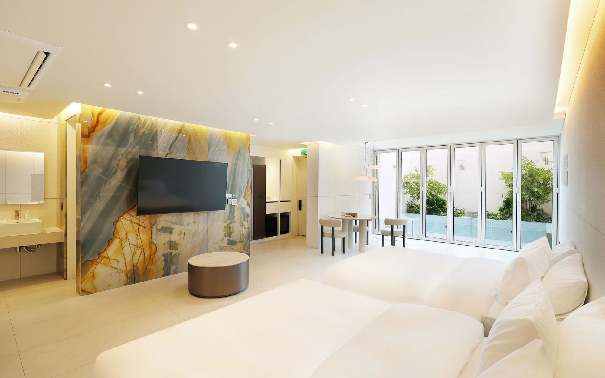 Una habitación de hotel moderna con una cama grande, un televisor de pantalla plana, una pared decorativa de mármol, un área de comedor con mesa y sillas, y un balcón con plantas.